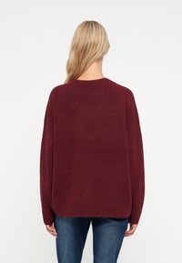 Burgundy oversized tröja med ribbat nackband, långa ärmar och en mjuk, strukturerad yta, parat med blå jeans.