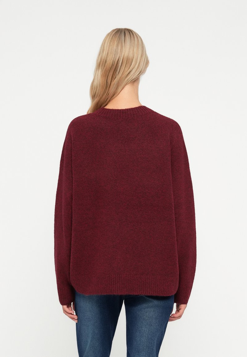 Sudadera oversized burdeos con cuello acanalado, mangas largas y un acabado suave y texturizado, combinada con vaqueros azules.