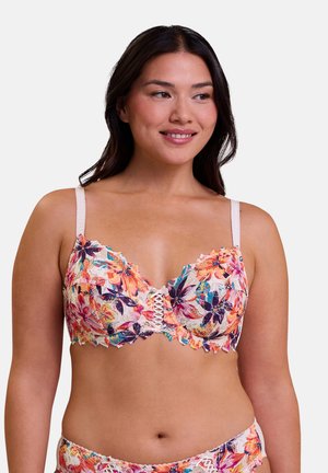 Femme portant un soutien-gorge en dentelle fleurie et un sous-vêtement assorti, souriante et regardant légèrement sur le côté sur un fond uni.