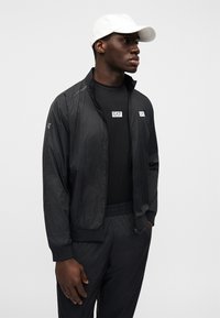 Μαύρο μπουφάν με φερμουάρ και καρό μοτίβο, με δύο μπαλώματα λογότυπου EA7. Φορεμένο πάνω από μαύρο πουκάμισο, συνδυασμένο με μαύρο παντελ όνι και λευκό καπέλο.