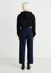 Crna skraćena dukserica s kapuljačom, nosi se preko tamnoplave široke traperice. Crne gležnjače upotpunjuju outfit. Vidljiva teksturirana tkanina.