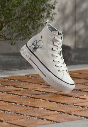 Weißer High-Top-Sneaker mit schwarzer und roter Cartoon-Grafik und Schnürsenkeln, schwebt über hölzernen Fliesen im Freien in der Nähe von grünem Laub und einer Betonwand.