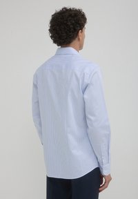 Camicia di cotone azzurra chiara con strisce verticali bianche, maniche lunghe e colletto classico. Texture liscia con polsini bottonati e orlo curvo.