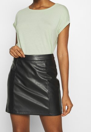 Persoon die een lichtgroene top met korte mouwen draagt, ingestopt in een zwarte leren minirok met hoge taille, staand tegen een effen achtergrond.