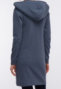 Person iført en lang, mørkeblå hættetrøje kjole, vist fra ryggen, stående mod en ensfarvet lys baggrund.