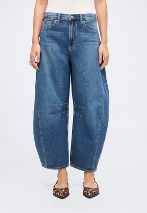 Γυναίκα που φοράει φαρδιά, άνετα μπλε τζιν με ορατές ραφές και μυτερές μπαλαρίνες με λεοπάρ μοτίβο, τα χέρια της ακουμπούν στα πλάγια.