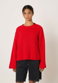 Pull en maille côtelée rouge avec un col rond et des manches larges, porté sur un short en denim noir. Le tissu texturé donne une apparence chaleureuse.