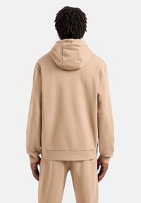 Felpa beige con cappuccio, dalla vestibilità ampia, texture morbida e polsini elasticizzati. Presenta una tasca a marsupio e un lacci regolabili sul cappuccio.