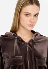 Sudadera con capucha de terciopelo marrón, con cremallera frontal, dos bolsillos en el pecho y acentos de metal. Textura suave, diseño ajustado.