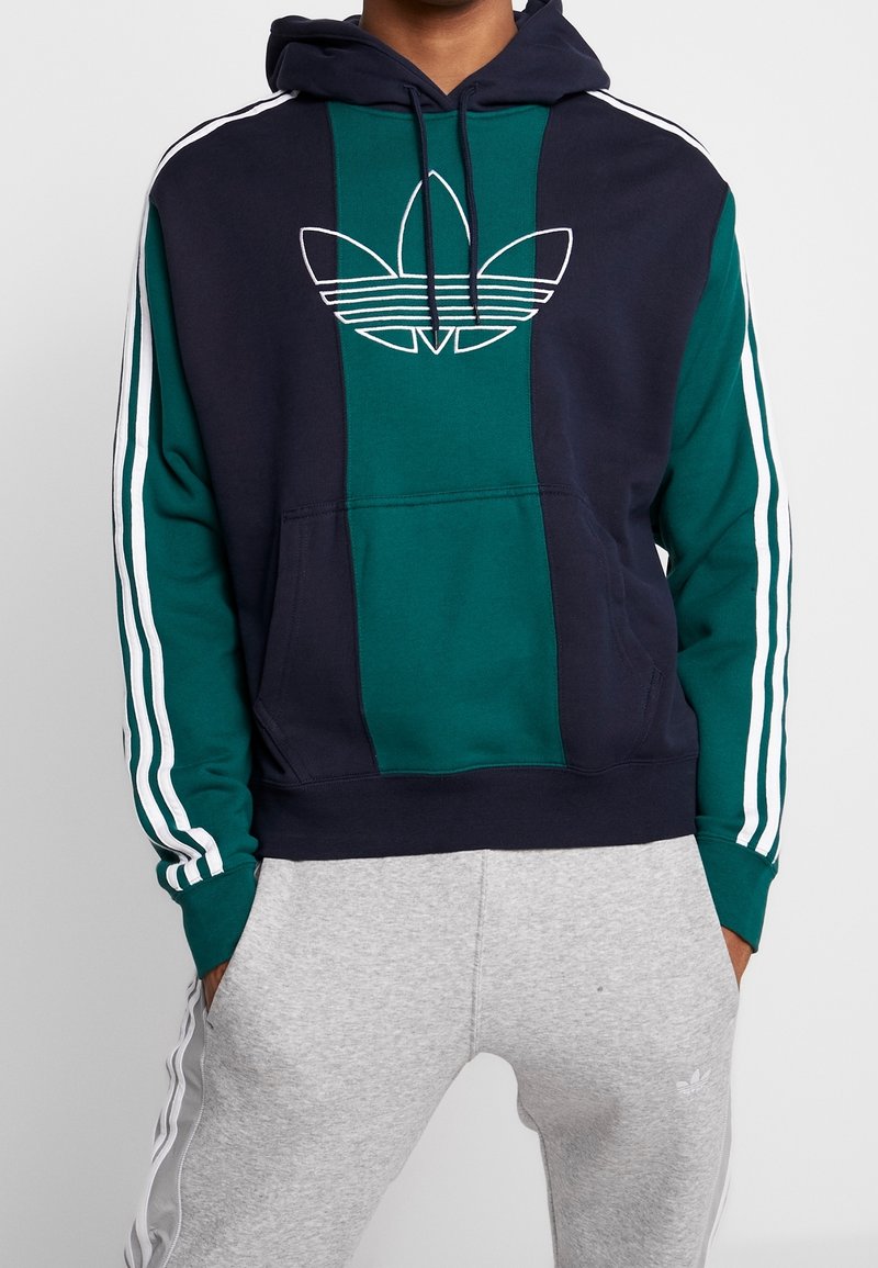 Sweatshirt à capuche avec un devant bleu marine et un panneau teal, logo blanc et rayures horizontales sur les manches. Le matériau semble doux et décontracté.