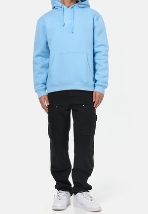 Sudadera con capucha de color azul claro con un bolsillo frontal, combinada con pantalones cargo negros que presentan una textura desgastada y zapatillas blancas.