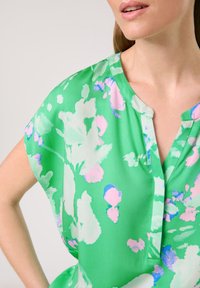 Grüne Blumenbluse mit kurzen Ärmeln, ausgestattet mit einem V-Ausschnitt und aus einem weichen, leichten Stoff. Rosa- und blaue Akzente verstärken das helle Design.