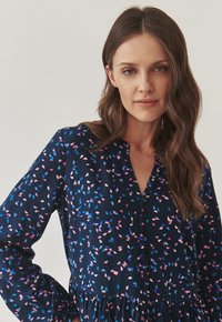 Donker marineblauwe jurk met een knoopsluiting en lange mouwen, versierd met een kleurrijk bladerenpatroon in roze en blauw, met een uitlopende zoom.