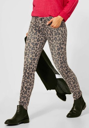 Vrouw draagt een broek met luipaardprint, zwarte enkellaarzen, rode trui, houdt een zwarte jas vast terwijl ze loopt.