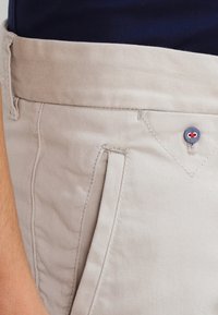 Beige bavlněné kalhoty s hladkou texturou, mají boční kapsy a knoflík s modrým základem a červeným akcentem na pase.