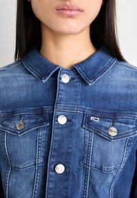 Denim jacket in dark blue with a button-up front, classic collar, two chest pockets, and metal accents. Subtle fading across the fabric.