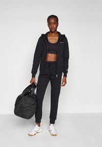 Schwarzes Sportoutfit bestehend aus einem Reißverschluss-Hoodie, einem Tanktop und Jogginghosen, kombiniert mit weißen Turnschuhen und einer schwarzen Sporttasche.