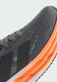 Scarpa sportiva grigia con tomaia texturizzata, strisce nere e suola intermedia arancione. Presenta lacci piatta e un'area del tallone rinforzata.