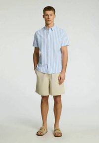 Camisa de botão de mangas curtas em azul claro e branco, combinada com calções bege e sandálias castanhas. Tecido suave e ajuste descontraído.