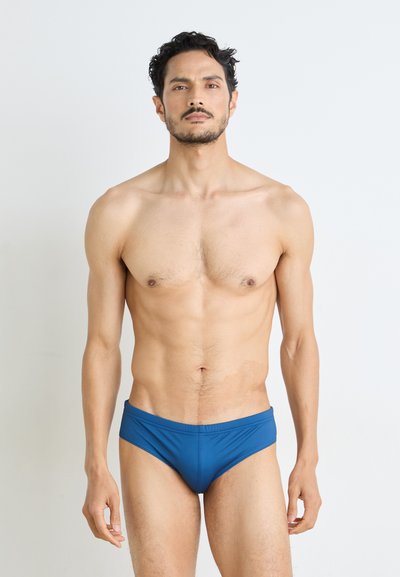 Blaue Badebriefe aus dehnbarem Material, mit einem tief sitzenden Design und glatter Textur, präsentiert von einem männlichen Model.