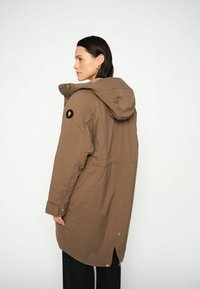 Parka marrone isolato con cappuccio, dotata di un patch con logo sulla manica e dettagli in oro, progettata con una vestibilità leggermente affusolata.