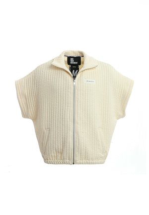 Joven con dreadlocks que lleva un chaleco acolchado cremado con cremallera, pantalones cortos beige, calcetines blancos con estampado y zapatos sin cordones texturizados, de pie con la mano cerca de la cara.