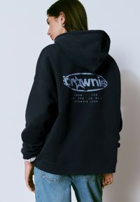 Sudadera negra con un gran gráfico en la parte posterior que presenta la palabra "brownie" y un diseño de estrellas. Tela suave, corte holgado y bolsillo tipo canguro.