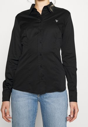 Kvinde iført en tætsiddende sort skjorte med knapper og krave samt lyseblå jeans, stående mod en ensfarvet hvid baggrund.
