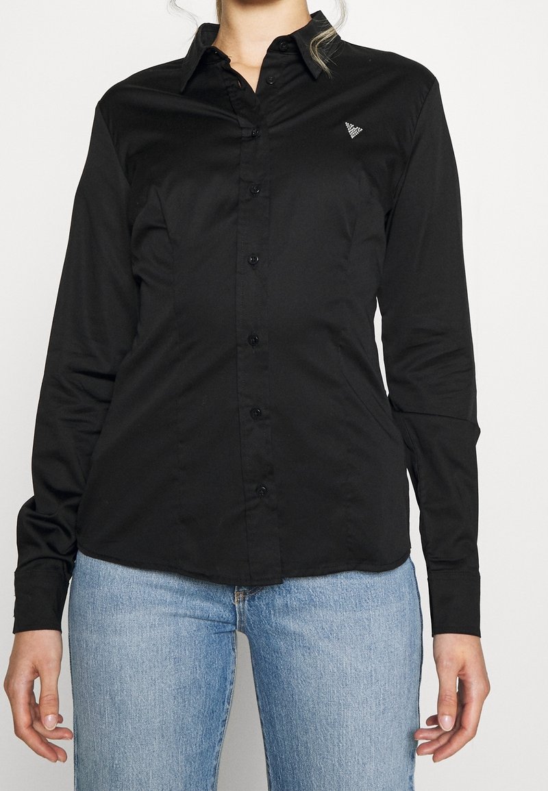 Femme portant une chemise noire cintrée à boutons avec col et un jean bleu clair, debout devant un fond blanc uni.