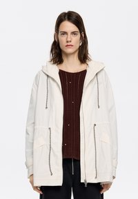 Hellbeige Kapuzenjacke mit Kordelzügen, Fronttaschen und Reißverschluss, über einem bordeauxfarbenen strukturierten Pullover getragen.