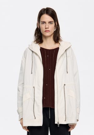Giacca cappuccio beige chiaro con coulisse, tasche frontali e chiusura con zip, indossata sopra un maglione a trama bordeaux.