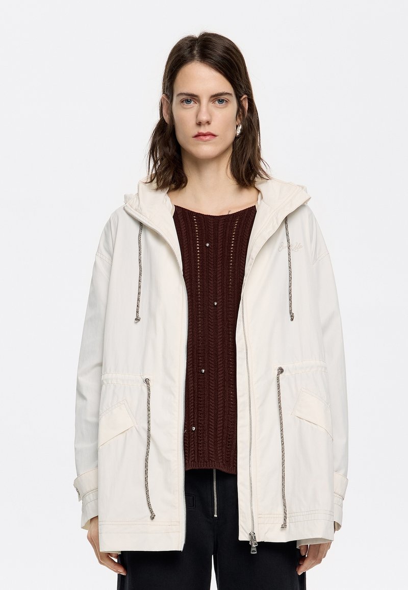 Hellbeige Kapuzenjacke mit Kordelzügen, Fronttaschen und Reißverschluss, über einem bordeauxfarbenen strukturierten Pullover getragen.