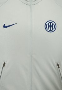 Svetlo šedá zipová bunda so hladkou textúrou, s námorníckou modrou značkou Nike a logom Inter Milána na hrudi, plus bočné zipsové vrecká.
