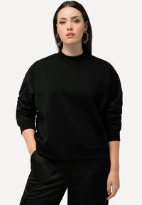 Sudadera negra de gran tamaño fabricada con un tejido suave, que presenta mangas largas y puños acanalados. Combinada con pantalones negros, creando un look monocromático.