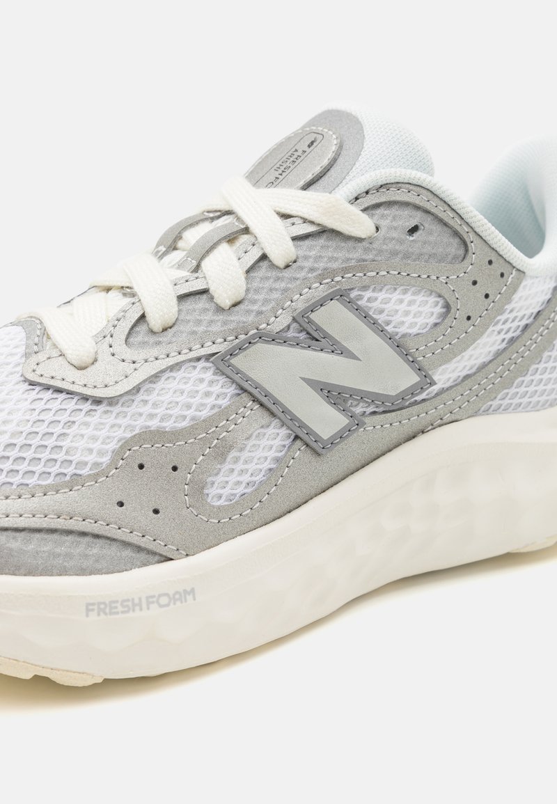Zapatilla deportiva gris y blanca con materiales de malla y gamuza, que cuenta con una suela de espuma gruesa y un prominente logo "N". Los cordones son de color blanco sucio.