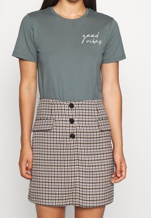 Vrouw die een grijs T-shirt met de tekst "good vibes" draagt, ingestopt in een hooggetailleerde beige en zwarte pied-de-poule rok met knopen aan de voorkant en zakken.