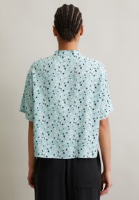 Short-sleeve, light blue shirt featuring a floral and abstract pattern in black, white, and green. Relaxed fit with a subtle collar design.