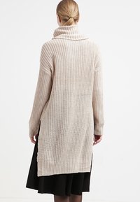 Kobieta z blond włosami w kucyku, ubrana w beżowy gruby sweter o długim kroju i czarną spódnicę, widziana z tyłu na jednolitym tle.