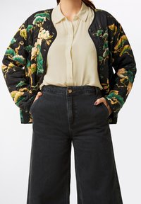 Quilted black jacket with floral patterns in green, yellow, and ivory, worn over a light beige blouse and high-waisted black denim pants.