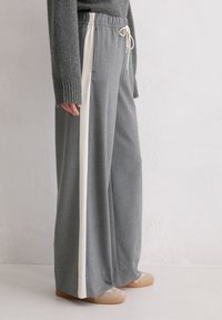 Osoba ubrana w szare szerokie spodnie z białymi paskami po bokach, w połączeniu z szarym swetrem dzianinowym i beżowymi butami typu slip-on.