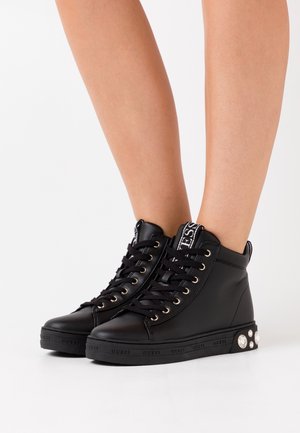 Černé členkové tenisky z umělé kůže mají zlaté očka, černé tkaničky, logo na jazyku a ozdobné kamínky na podrážce.