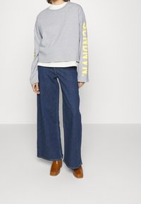 Gray cropped sweatshirt with yellow lettering, paired with wide-leg blue denim jeans and brown block-heel shoes.