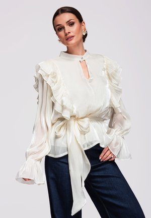 Femme portant un chemisier blanc transparent avec des épaules et des poignets à volants, noué à la taille, associé à un jean bleu foncé, posant devant un fond blanc.