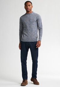 Hombre de pie que lleva un suéter de punto azul grisáceo, jeans oscuros y botas marrones con cordones, frente a un fondo claro y liso.