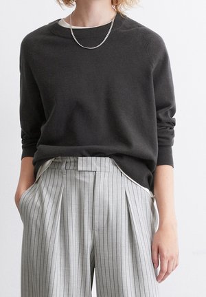 Person iført en mørkegrå langærmet sweater, sølvkæde halskæde og højtaljede lysegrå bukser med brede ben og tynde striber.