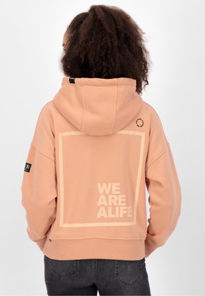 Personne aux cheveux bouclés portant un sweat à capuche pêche avec le texte "WE ARE ALIFE" encadré au dos, porté avec un jean foncé, vue de dos.