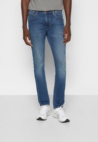 Pantalones vaqueros de mezclilla azul con un corte ajustado, desvanecimiento sutil, cinco bolsillos y herrajes de latón. Llevados con una camiseta gris y zapatillas blancas.