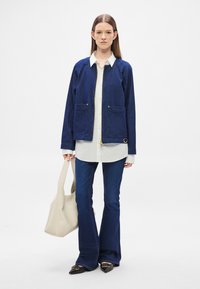 Τζιν μπουφάν σε σκούρο μπλε, με μπροστινό φερμουάρ, δύο μεγάλα τσέπες και στρογγυλεμένο γιακά. Συνδυασμένο με λευκό πουκάμισο, φαρδιά τζιν και μπεζ τσάντα.