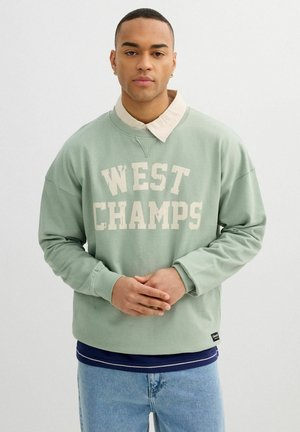 Joven con el pelo corto, viste una sudadera verde claro con la inscripción "WEST CHAMPS", camisa beige con cuello y vaqueros azul claro, de pie con las manos juntas.