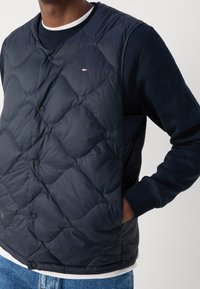 Marineblaue gesteppte Weste mit runden Rautensteppungen, ausgestattet mit schwarzen Druckknöpfen und einem kleinen Logo, über einem marineblauen Langarmshirt getragen.